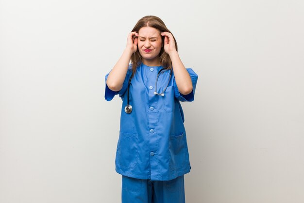 Young nurse woman against a white wall touching temples and having headache.