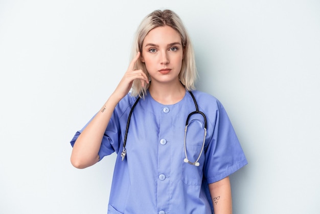 Молодая женщина медсестры изолирована на синем фоне указывая храм пальцем, думая, сосредоточился на задаче.