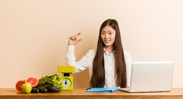 Young nutritionist chinese woman working with her laptop holding something little with forefingers, smiling and confident.