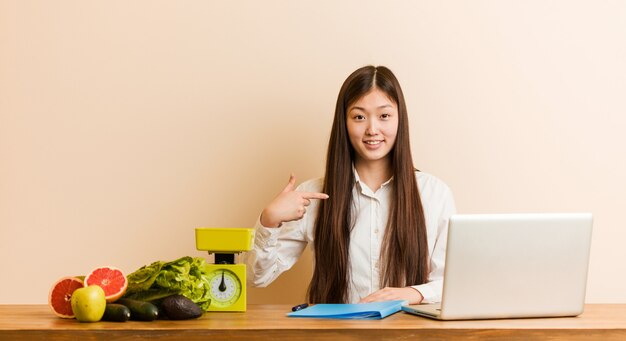 誇りと自信を持って、シャツのコピースペースを手で指している彼女のラップトップの人と一緒に働いている若い栄養士の中国人女性