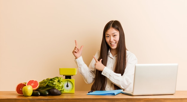 Young nutritionist chinese woman working with her laptop pointing with forefingers to a copy space, expressing excitement and desire.
