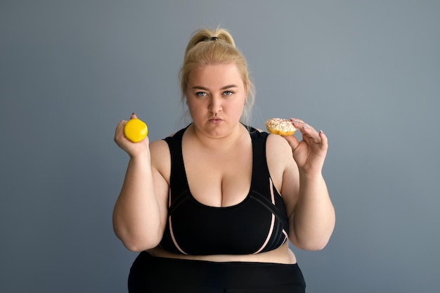 A young obese woman holds dumbbells and a bagel in her hand with a hungry and angry look