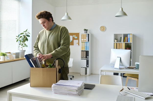 Young office worker standing at his workplace and packing his things in box after layoff