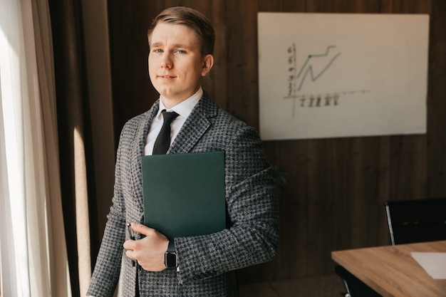 A young office worker with fair skin holds a folder with documents in his hands, stands in his office. The man works in the office.