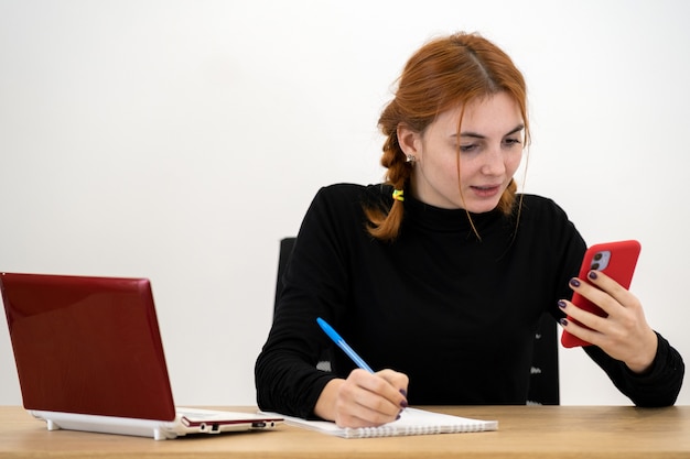 Young office worker woman talking on a cell phone sitting behind working desk with laptop computer and notebook.