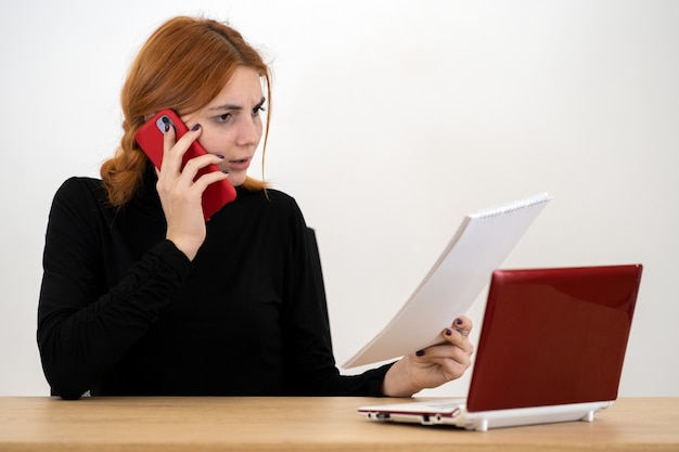 Foto giovane donna di impiegato che parla su un telefono cellulare che si siede dietro lo scrittorio funzionante con il computer portatile ed il taccuino.