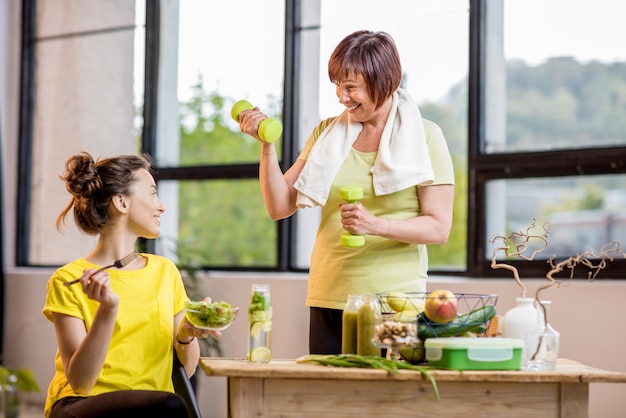 ダンベルで運動し、窓の背景に屋内で健康的な食べ物を食べる若い女性と年配の女性