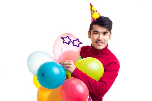 Young optimistic man with celebrating hat holding many colored balloons and card stick of glasses