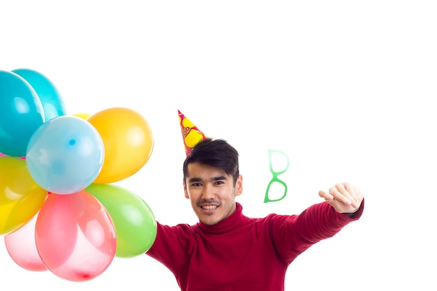 Young optimistic man with celebrating hat holding many colored balloons and card stick of glasses