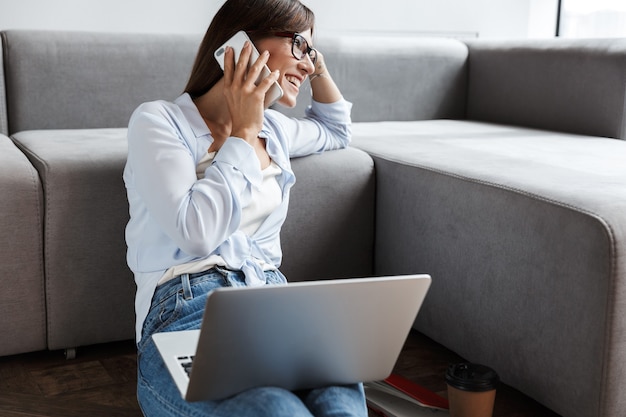 молодая оптимистичная довольная деловая женщина в помещении у себя дома на полу возле дивана с помощью портативного компьютера разговаривает по мобильному телефону.