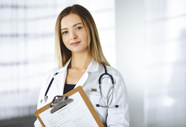 Young optimistic woman-doctor is holding a clipboard in her hands, while standing in a clinic. Portrait of friendly female physician with a stethoscope. Perfect medical service in a hospital. Medicine