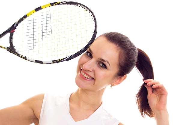 Foto giovane donna ottimista in camicia sportiva bianca con coda di cavallo scura che tiene una racchetta da tennis in studio