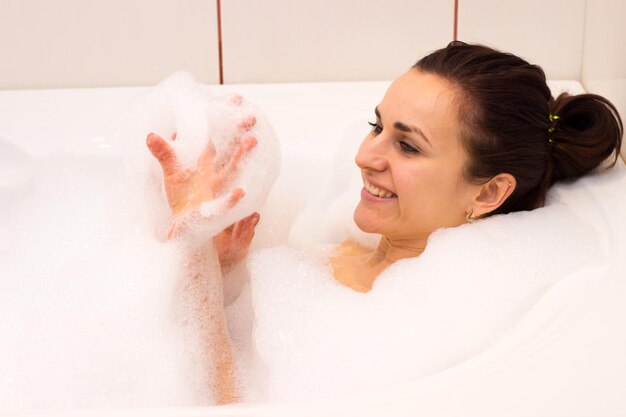 Young optimistic woman with bundle on her head lying in the bath full of foam