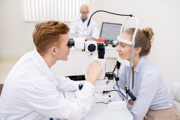 Молодой окулист в белом халате смотрит через линзу оборудования для проверки зрения перед пациенткой