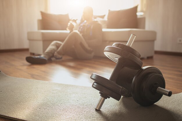 Young ordinary man go in for sport at home. Regular guy sit at sofa on floor after hard workout. Has rest and drink water. Blurred sunny background. Pair of dumbbells lying on mat in front.