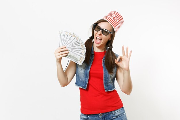 Young overjoyed pretty woman in 3d glasses with bucket for popcorn on head watching movie film, holding bundle of dollars, cash money, showing OK sign isolated on white background. Emotions in cinema.