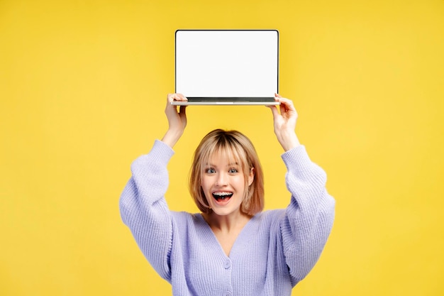 Young overjoyed woman holding laptop computer showing blank monitor isolated on yellow background
