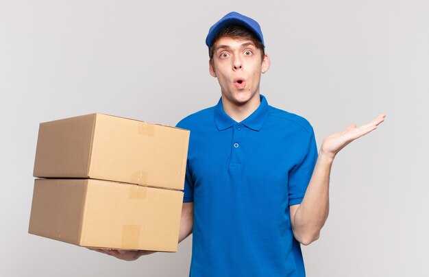 Young package deliver boy looking surprised and shocked, with jaw dropped holding an object with an open hand on the side