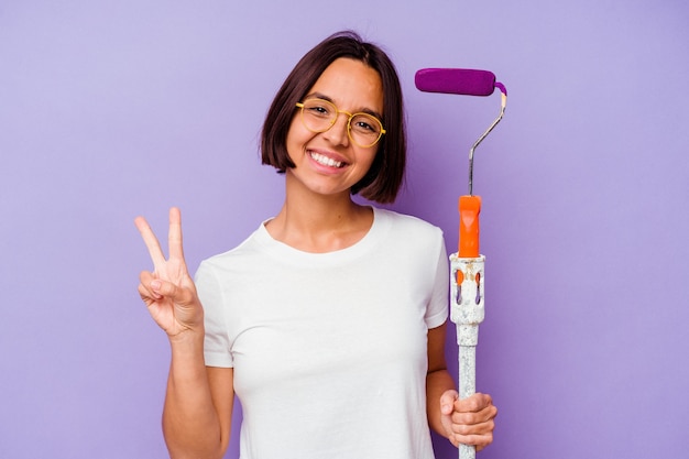 Young painter mixed race woman holding a paint stick isolated on purple background joyful and carefree showing a peace symbol with fingers.