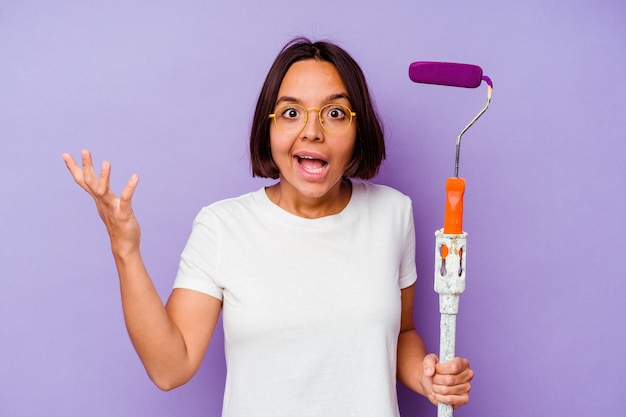 Young painter mixed race woman holding a paint stick isolated on purple background receiving a pleasant surprise, excited and raising hands.