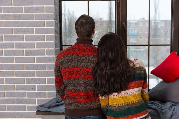 따뜻한 니트 스웨터를 입은 젊은 쌍은 베개와 담요가 있는 창턱 근처에 서서 창 밖을 내다보고 있다