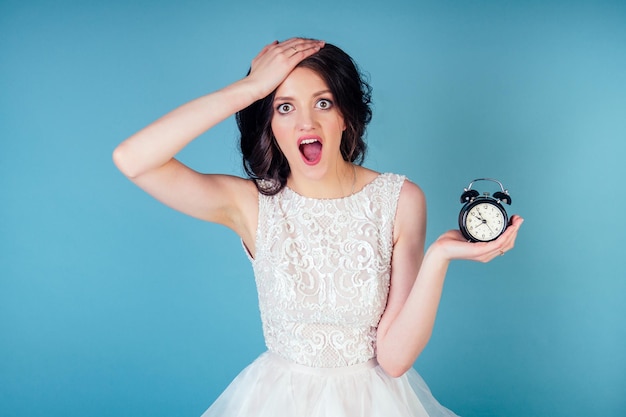 Young and panic beautiful woman bride with makeup in white wedding dress holds an alarm clock in hand and screams concept of being late for a wedding ceremony date I am late to my wedding