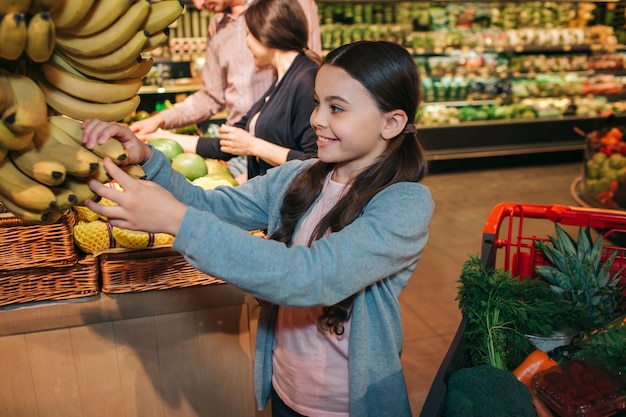 Young parents and daughter in grocery store. Child touch yellow bananas and smile. She is happy. Man and woman stand behind and choose fruit.