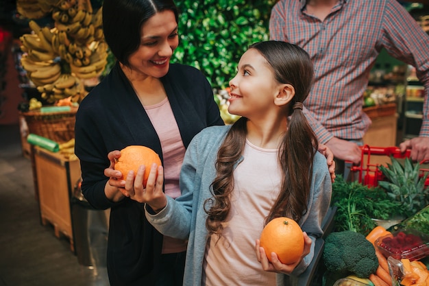 Young parents and daughter in grocery store. Little girl look at parents and smile. She hold oranges in hands. Father stand behind.