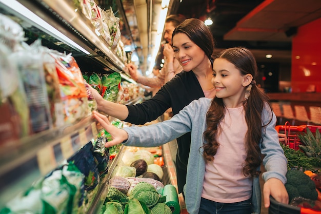 Young parents and daughter in grocery store. Positive woman and child pick up together salad. Father stand behind.