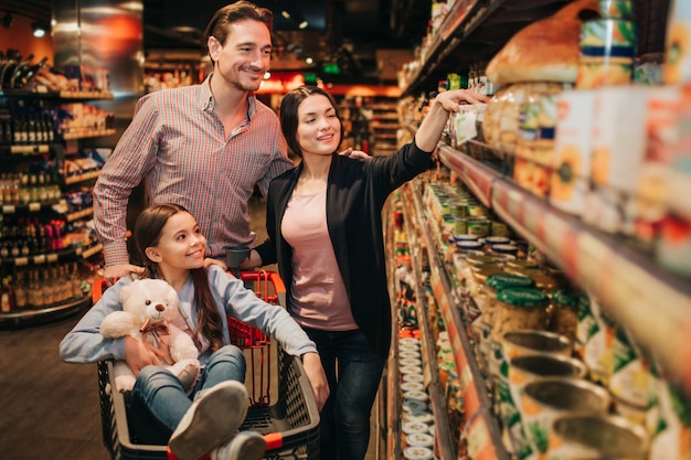 Young parents and daughter in grocery store. They pick up conservation together. Girl sit in grocery trolley woth bear toy.