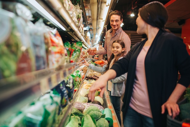 Young parents and daughter in grocery store. Woman look back and point down. Father and daughter look at her.