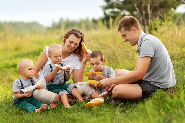 Young parents and their children sit on the grass and eat melon