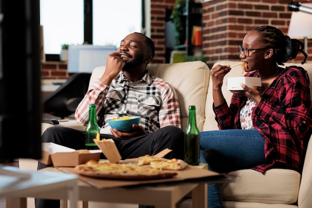 Young partners enjoying fast food delivery with movie on tv, having fun with leisure activity. Eating takeaway meal with noodles, pizza and chips, drinking beer from bottle. Watching television.