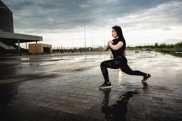 Young passen vrouw in sportkleding doen lunges. De vrouw leidt in openlucht na regen op. Fitness, training, sport buitenshuis concept.