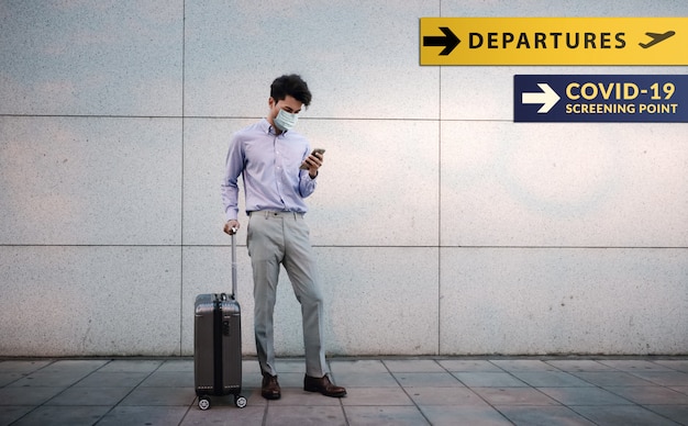 Giovane uomo d'affari del passeggero che indossa maschera chirurgica. utilizzando smartphone. in piedi con i bagagli in aeroporto.