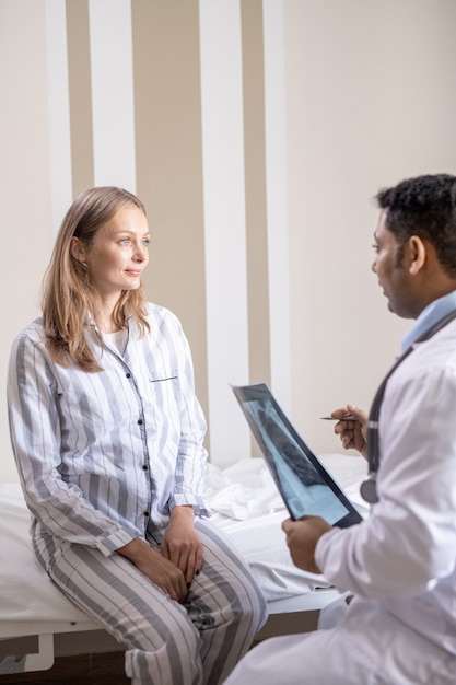 Young patient of covid hospital sitting on bed in front of her doctor during discussion of x-ray results, medical prescriptions and recommendations