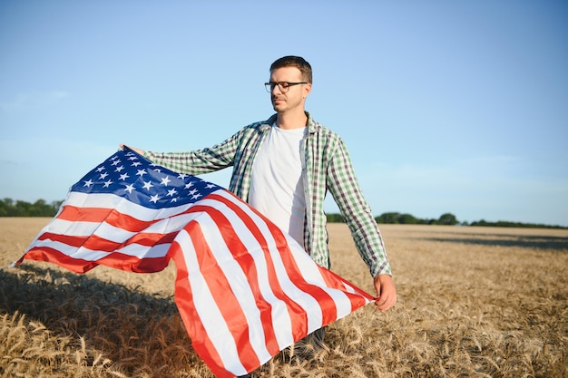 Il giovane agricoltore patriottico si trova tra il nuovo raccolto ragazzo che cammina con la bandiera americana sul campo di grano che celebra il giorno dell'indipendenza nazionale 4 luglio concetto