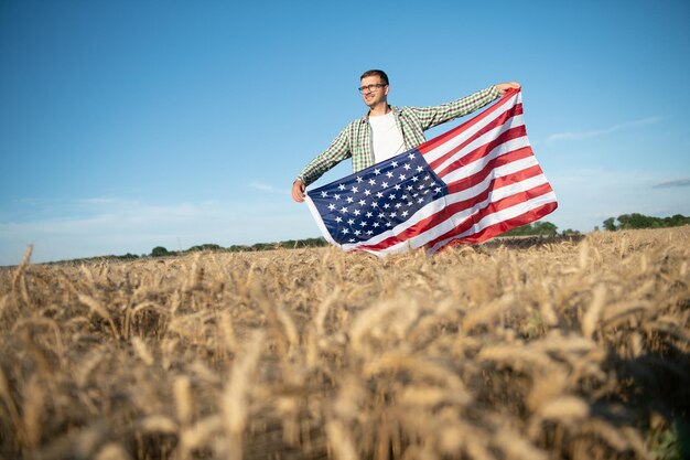 Young patriotic farmer stands among new harvest. Boy walking with the american flag on the wheat field celebrating national independence day. 4th of July concept.