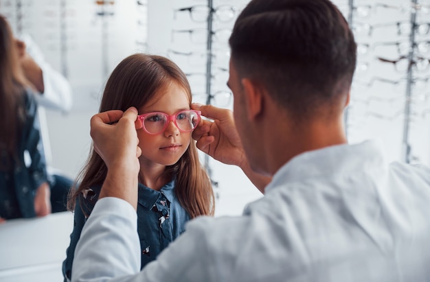 白衣を着た若い小児科医は、小さな女の子のための新しい眼鏡を手に入れるのを手伝います。