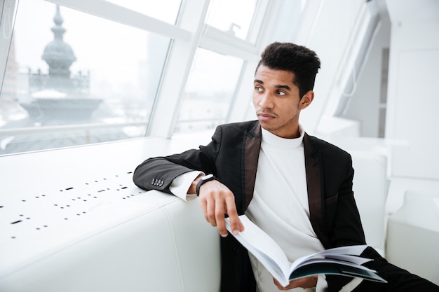 Young Pensive african business man in black suit sitting on white sofa and holding journal in office