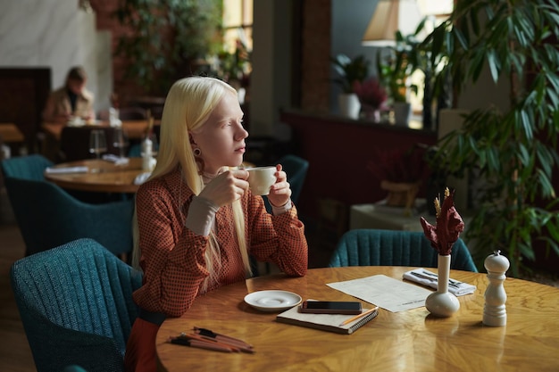 Молодая задумчивая женщина-альбинос с чашкой чая отдыхает за столиком в кафе