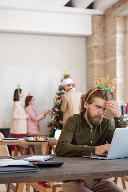 Young pensive bearded businessman with Christmas headband looking at laptop display while sitting by desk, brainstorming and networking
