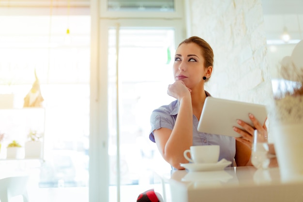 カフェで休憩中の若い物思いにふける女性。彼女はデジタルタブレットを使用してコーヒーを飲んでいます。