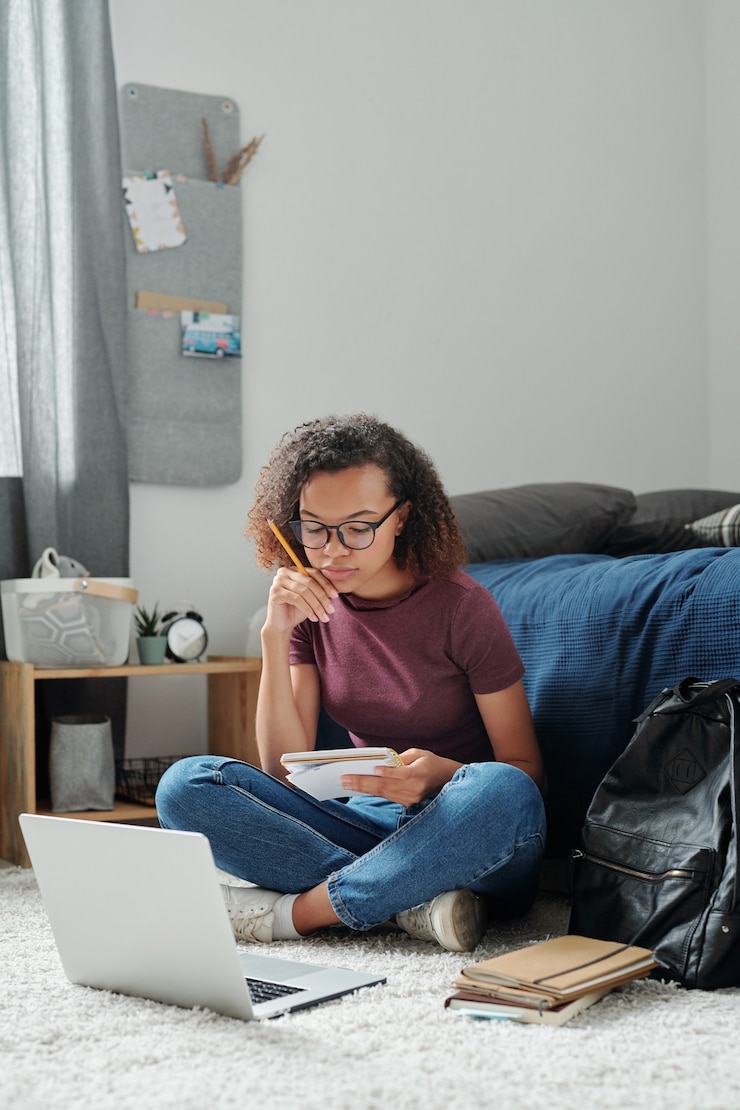 Photo young pensive cross-legged female student sitting on the floor by bed in front of laptop and looking through her notes in copybook in bedroom