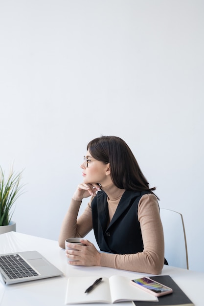 Young pensive female analyst with drink thinking of how to deal with financial situation of company in front of laptop