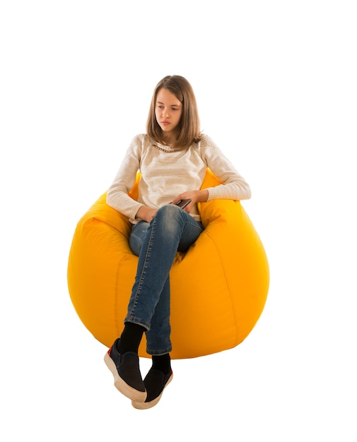 Young pensive girl holding the tablet while sitting on yellow beanbag isolated on white