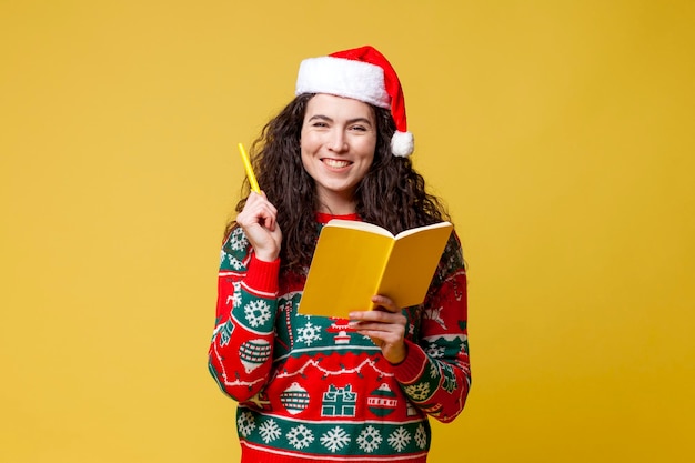 Young pensive girl in New Year's clothes and santa hat holds book and plans on yellow background