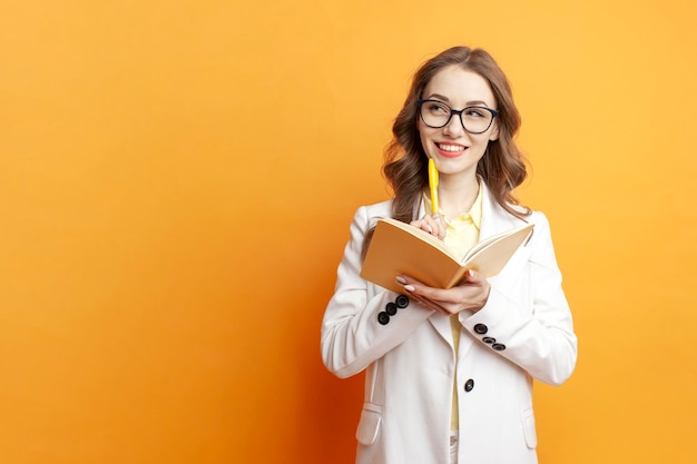 Young pensive girl student in glasses and white suit writes in yellow notebook and dreams