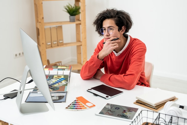 Молодой задумчивый веб-дизайнер в очках, глядя на экран компьютера, работая над новым логотипом дома