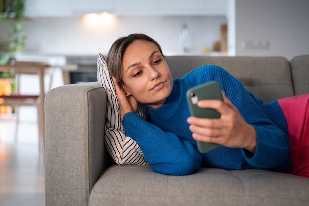 Молодая задумчивая женщина в синем свитере читает плохие новости от парня в социальных сетях по телефону
